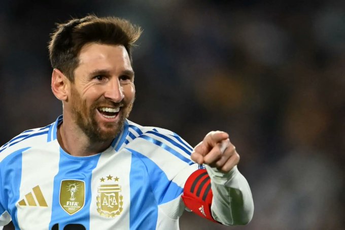 Lionel Messi in Argentina national team blue and white jersey with Adidas stripes and sponsor patches stands on soccer field smiling and pointing forward arm extended crowd blurred in background stadium setting.
