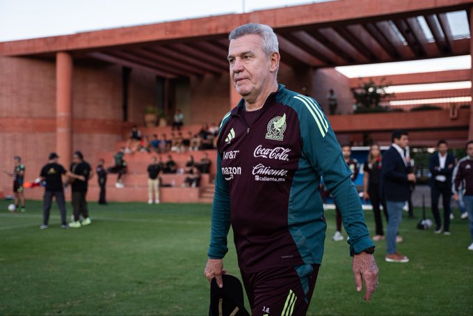 Older man with gray hair and beard wearing green and maroon Adidas jersey with Pumas and sponsor logos walks on green soccer field holding black cap, red brick stadium structure and seating in background with scattered spectators in casual attire standing or sitting nearby under open sky.
