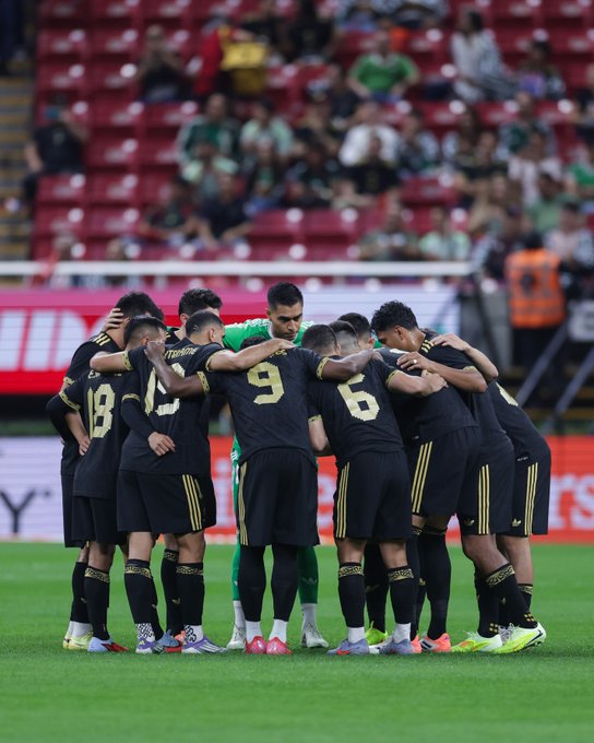 Group of Mexican national soccer team players in black and gold jerseys forming a huddle on green soccer field with stadium seating and spectators in background red and green seats visible some players with hands on heads coach in green jacket center.