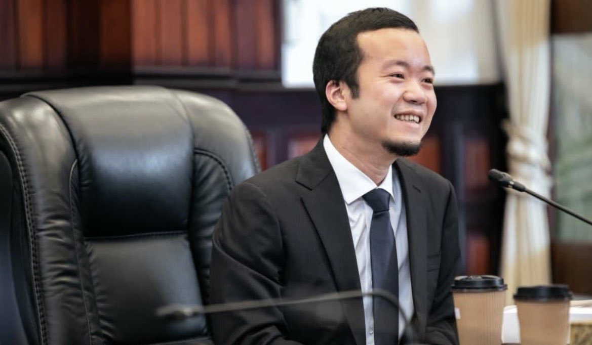 A man in a dark suit with white shirt and tie sits smiling in a large leather chair at a wooden desk in a formal room with bookshelves and curtains behind him. Two microphones are positioned in front of him on the desk. Two white disposable coffee cups with lids sit on the desk to his right.