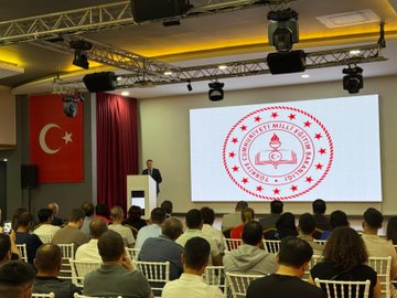 First image shows a conference room setup with lighting rigs, stage, large screen displaying a red circular emblem with Turkish text encircled by stars and flames, Turkish flags on stands, red curtains, and audience seated in white chairs facing the stage with a male speaker at podium. Second image depicts a large group of men and women in formal suits, dresses, and traditional attire standing in rows on a wooden floor with yellow wall panels, chairs, and a pedestal in a modern hall. Third image features a woman in professional attire speaking on stage in front of a large MEB logo with flame and house symbol, red curtains, portrait of a man on wall, audience seated in burgundy chairs including some in headscarves. Fourth image illustrates two men speaking on stage with a screen showing a heart and house emblem, Turkish flag, portrait on wall, audience in red chairs in a hall with orange lighting and patterned walls.