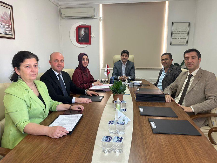 A group of six formally dressed professionals sits around a wooden conference table in an office with beige walls and a window. The individuals include men in suits and women in blouses and hijabs, with one man at the head position. Turkish flags stand on the table, alongside documents, water glasses, a potted plant, and notepads. A framed certificate hangs on the wall, and a red crescent symbol is visible on another wall.
