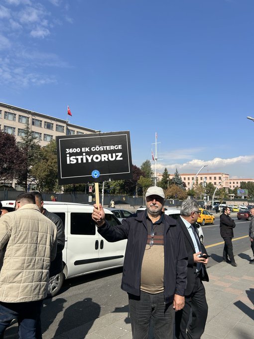 First image shows a man in a cap and jacket holding a black sign reading 3600 EK Gosterge Istiyoruz near a government building with Turkish flag, surrounded by other people and vehicles on a sunny day. Second image depicts several men in suits holding blue and red signs with messages like MEMUR EMEKLI ARTISI YOK SAĞLIK EMEĞİ and ÖĞRETIM EMEĞİ, standing in an open area with trees and flags. Third image features a man in a blue jacket holding a white sign reading EK ZAM VE REFAH PAYI ISTIYORUZ near trees and a speaker setup in a grassy area. Fourth image shows a group including a man in a red vest holding a sign reading ENFLASYONA KARŞI BİRLİK OL and another with MEMUR EMEKLİSİ, near trees and a speaker on grass.