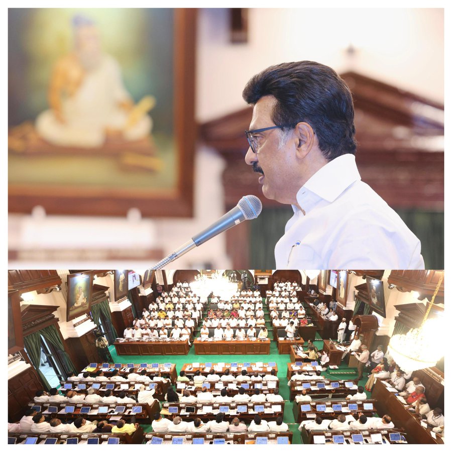 A framed portrait of a revered figure in traditional attire seated with hands in gesture inside an ornate hall, and a man in white shirt and glasses speaking into a microphone from a podium in the foreground. Below, a large green-carpeted assembly hall filled with rows of seated individuals in formal attire, some wearing yellow shirts, under bright lighting with wooden architecture and multiple microphones visible.