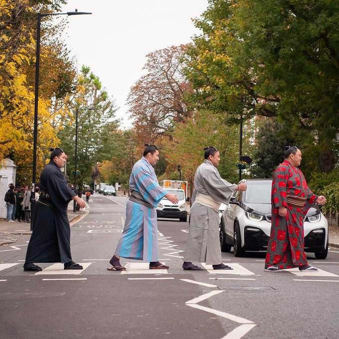 Four sumo wrestlers dressed in traditional Japanese robes of varying colors including black, blue, gray, and red floral patterns walk across a zebra crossing on a residential street. They are positioned in a line mimicking a famous album cover pose with one in front leading and another holding a small camera. Surrounding elements include autumn foliage on trees, a white BMW i3 electric car parked nearby, a delivery van in the background, street lamps, and a few pedestrians observing from the sidewalk under a cloudy sky.
