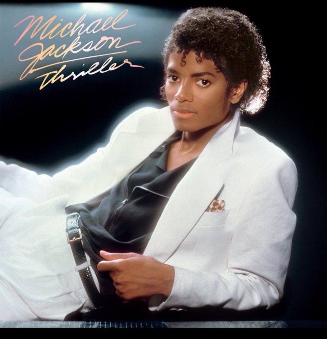 Michael Jackson with curly black hair sits leaning forward on a dark background, wearing a white suit jacket over a black shirt and pants with a belt, one hand gesturing with fingers extended, gold lettering above reads Michael Jackson in cursive and Thriller below in block letters.