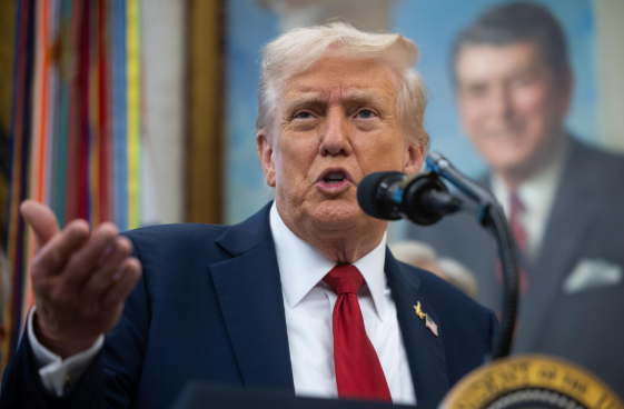 Donald Trump stands at a wooden podium with a microphone, wearing a dark suit, white shirt, red tie, and American flag pin, gesturing with his right hand while speaking, with a blurred background of colorful flags and a large portrait of Ronald Reagan on the wall behind him.