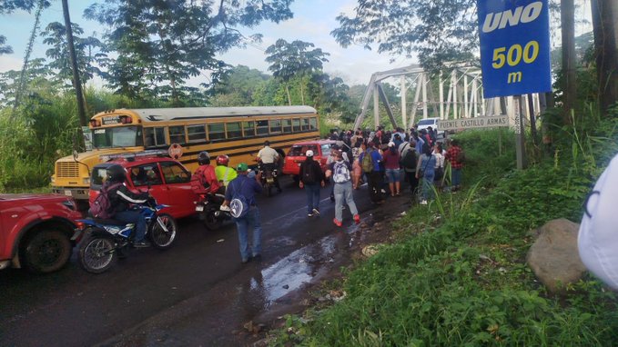 First image shows a yellow school bus parked near a bridge with a sign reading UNO 500 M surrounded by trees and wet road a crowd of people including students with backpacks standing around red vehicles and motorcycles on a rural road in a forested area. Second image depicts a white truck and red vehicles with a group of people including a man in white shirt and cap observing the bridge sign UNO 500 M with lush greenery and wet ground nearby. Third image features several officials in uniforms and casual attire including vests and hats standing on a dirt road near a white truck and yellow vehicle under trees with a blue flag visible in the background.