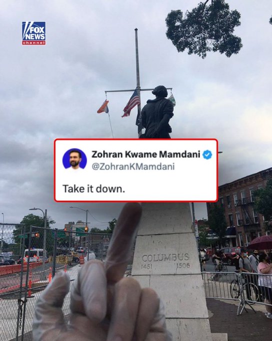 Urban street scene with a tall statue of Christopher Columbus on a pedestal inscribed 1492, surrounded by American and other flags on poles under a partly cloudy sky. A red-bordered tweet bubble from Zohran Kwame Mamdani displays text Take it down with a profile image of a man. A hand extends a middle finger toward the statue base. Background includes buildings, traffic lights, vehicles, and people.