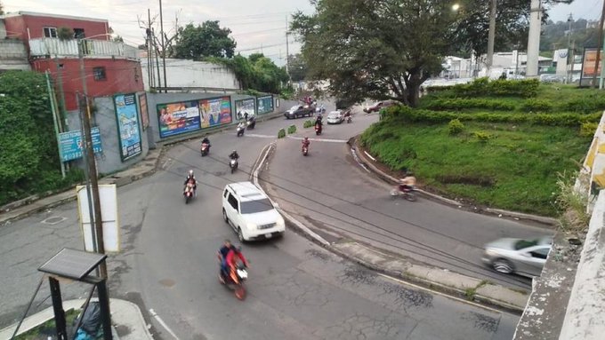 Curved urban road with multiple motorcycles and cars including a white SUV navigating smoothly, surrounded by greenery, billboards on a fence, buildings, trees, and utility poles in a residential area under overcast sky.