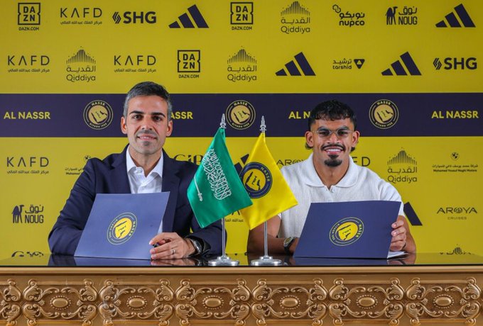 Two men seated at a table during a formal signing event one in suit holding blue folder with Al Nassr logo other in white polo holding yellow folder Saudi green flag and yellow flag on stands in front Al Nassr team emblem backdrop with sponsor logos including KAFD SHG Adidas DZN Qiddiya Nupco yellow background ornate golden table edge