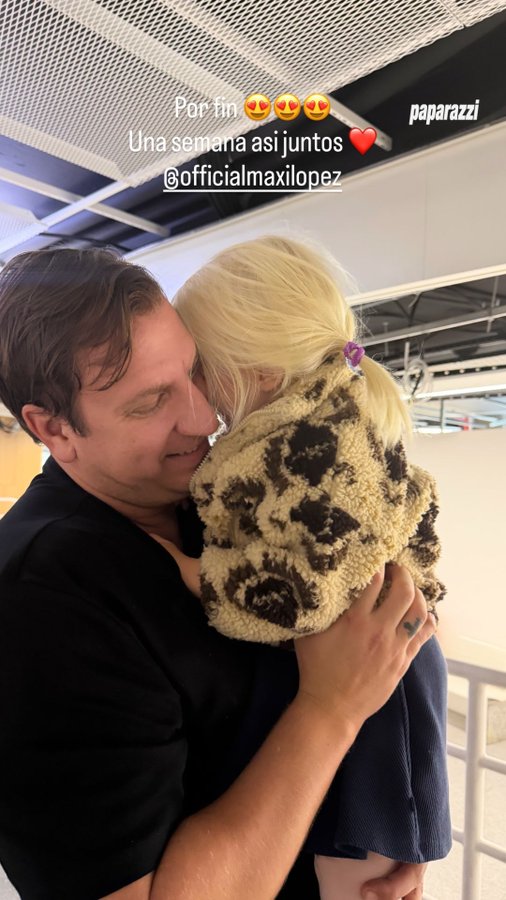 A man with short dark hair wearing a black shirt holds and smiles at a young blonde girl in a leopard-print outfit with pigtails, both in an indoor space with ceiling panels, tagged with Paparazzi watermark and official Instagram mention.