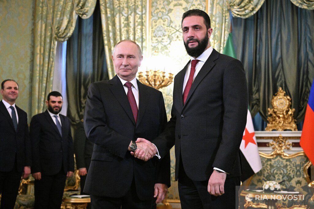 Vladimir Putin in a dark suit and red tie stands shaking hands with Bashar al-Assad in a dark suit and red tie inside a grand room with elaborate green drapery, ornate golden decorations, a chandelier, and flags of Russia and Syria visible on a table, while several men in dark suits with red ties stand nearby observing the handshake, with RIA Novosti watermark in the corner.