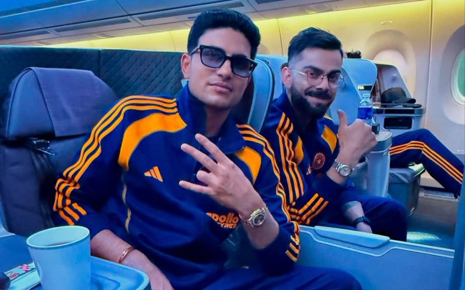 Two men, Virat Kohli and Shubman Gill, seated side by side in an airplane cabin with blue seats and overhead lighting. Both wear blue and orange Adidas tracksuits featuring sponsor logos on the chest. Virat Kohli on the left has short dark hair, wears dark sunglasses, and holds up a peace sign with his right hand. Shubman Gill on the right has short dark hair, wears clear glasses, and also makes a peace sign. A blue water bottle sits on the tray table in front of them, along with wristwatches on their left wrists and a bracelet on Gills right wrist. The background includes airplane windows and cabin interior.