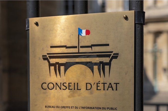 Brass plaque mounted on a black metal frame against a stone wall with barred windows in background, displaying the French tricolor flag above a silhouette of a bridge, text CONSEIL D'ÉTAT in large letters, and below it BUREAU DU GREFE ET DE L'INFORMATION DU PUBLIC in smaller letters.
