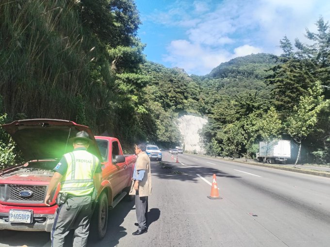 Two individuals in high-visibility vests stand beside a red pickup truck with its hood open on a roadside surrounded by lush green forested hills and a mountain in the background under a partly cloudy blue sky. The truck appears to have mechanical issues as tools and parts are visible near the engine. Orange traffic cones are placed for safety along the asphalt road with other vehicles including a white car and a truck passing by in the distance. The scene is set in a rural mountainous area with dense vegetation lining the highway.