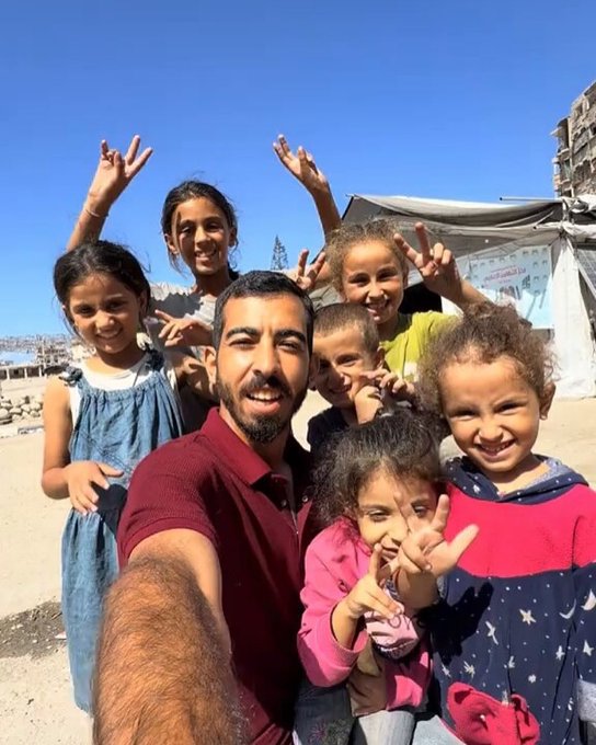 Group of diverse children and one adult man posing for selfie in sunny outdoor refugee camp setting with blue sky overhead several children wearing casual clothes like dresses hoodies and t-shirts making peace sign gestures with hands man in red polo shirt smiling while holding phone for photo tents and damaged buildings visible in background sandy ground underfoot