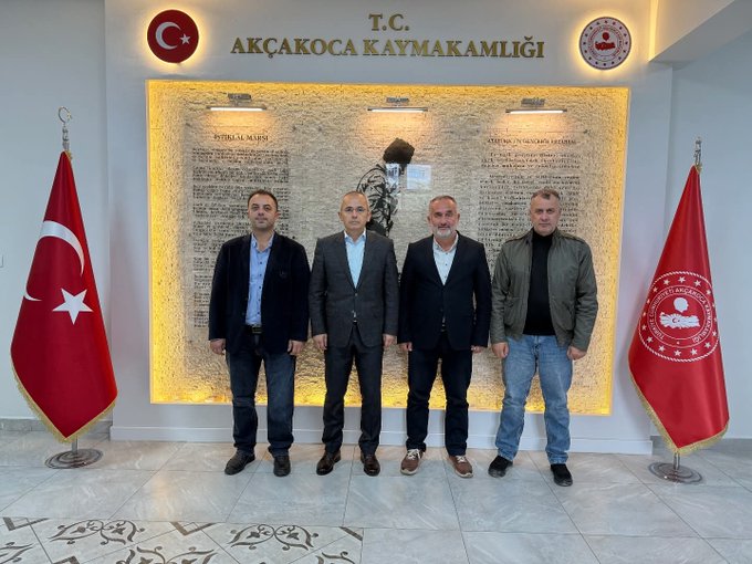 Group of four men standing in formal attire in front of a wall display at Akçakoca Kaymakamlığı office featuring Turkish flags on both sides illuminated panels with inscribed text and a central portrait of Mustafa Kemal Atatürk two additional Turkish flags and official emblems visible in the background.