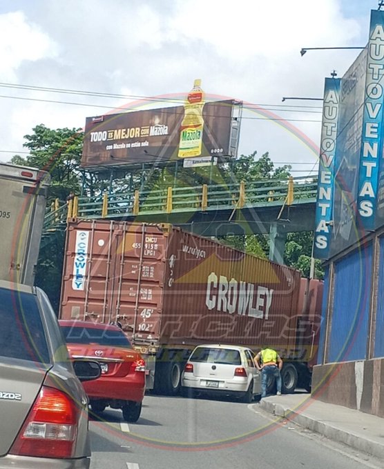 Urban street scene in Guatemala with a large yellow billboard advertising Modelo beer on the left side, a pedestrian overpass with yellow railings in the background, multiple cargo trucks including a brown Crowley truck with white lettering and license plate visible, several cars like a red sedan and white van in traffic, a worker in yellow safety gear near the truck, green trees lining the road, and blue buildings nearby.