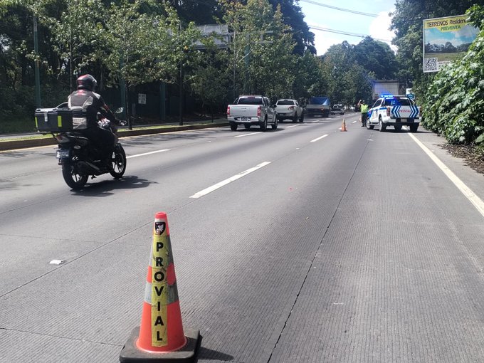 A motorcycle with a rider in uniform stops near orange and white traffic cones marked Provial on a multi-lane highway. A white pickup truck and police vehicle with blue and white stripes are parked nearby. Green trees line the roadside with a yellow sign visible in the background. Additional vehicles including a white truck are positioned along the road. The scene depicts roadside assistance for a stranded vehicle obstructing traffic.