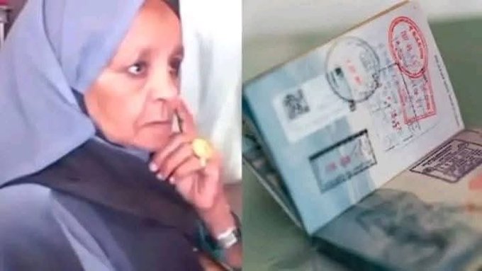 A woman wearing a light blue headscarf and dark clothing sits and holds an open passport booklet with her right hand, examining it closely while wearing a yellow ring on her finger and a watch on her wrist. The passport displays multiple red stamps, barcodes, and text pages visible on the spread. The background includes a plain surface like a table.