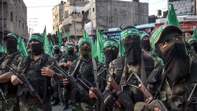 A group of armed men wearing green headbands and black masks stand in a line on a street in an urban area with buildings and Palestinian flags visible in the background, dressed in military-style camouflage uniforms and carrying rifles.