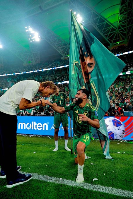 Players in green jerseys celebrate on a soccer field in a stadium, one holding a large green flag with a persons face printed on it, another player in white shirt and green pants raising his arm, stadium lights and seating visible in background, Molten sponsor logo on field.