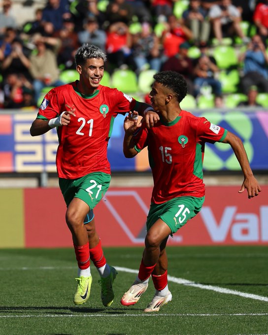 Dos jugadores de fútbol marroquíes con camisetas rojas y verdes numeradas 21 y 15 corren celebrando en un campo de césped verde con líneas blancas, fondo de estadio con asientos verdes y rojos ocupados por espectadores, publicidad en las barreras laterales y porterías visibles al fondo.
