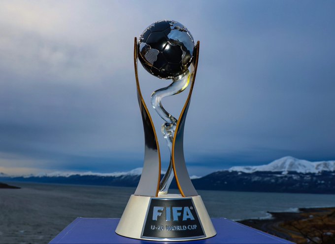 The FIFA U-20 World Cup trophy stands on a white base with the text FIFA U-20 World Cup engraved on it. The trophy features a black globe held by a twisted metallic structure with gold accents. It is placed on a purple surface outdoors against a backdrop of calm water, distant snowy mountains, and a partly cloudy blue sky.