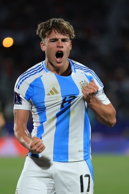 A young male soccer player with blonde hair wearing the Argentina national team jersey number 17 in blue and white stripes with gold accents stands on a stadium field at night under lights. He has his mouth open wide in a shout of celebration with his right fist clenched and raised while his left hand pulls at his jersey collar. The background shows a blurred stadium crowd and a yellow tennis ball visible in the air.