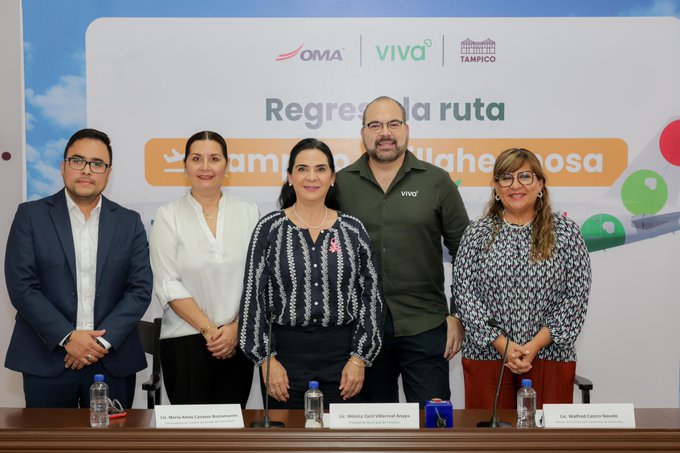 Group of five professionals standing behind a conference table during a press announcement. Leftmost man in suit and glasses, next woman in white blouse, then woman in striped blouse with red pin, man in green shirt with Viva Aerobus logo, and rightmost woman in patterned blouse with glasses. Table has water bottles and nameplates. Backdrop features Tamaulipas Viva branding with orange text Regresa la ruta Tampico-Villahermosa, airline logos, and colorful graphics including green and orange elements against a blue sky background.