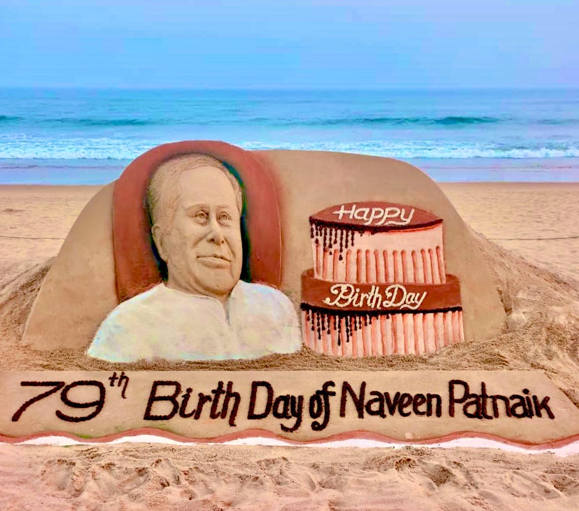 A detailed sand sculpture on a sandy beach with ocean waves in the background shows a realistic portrait of a man with short hair and glasses wearing a white kurta, positioned next to a three-tiered birthday cake sculpted from sand with dripping icing effects and the text Happy Birthday written on the top tier. The base of the sculpture includes the inscription 79th Birthday of Naveen Patnaik in sand. The artwork is created with fine details in the facial features and clothing folds, set against a clear blue sky.
