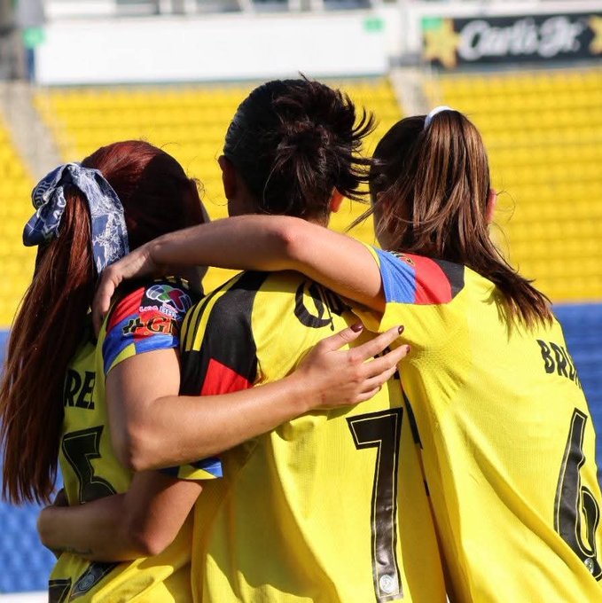 Three female soccer players stand back-to-back in a stadium, arms around each other in celebration. They wear yellow jerseys with black accents, one with number 7 and name BRE, another with 6. Red hair ties and ponytails visible, stadium seats in yellow and blue background.