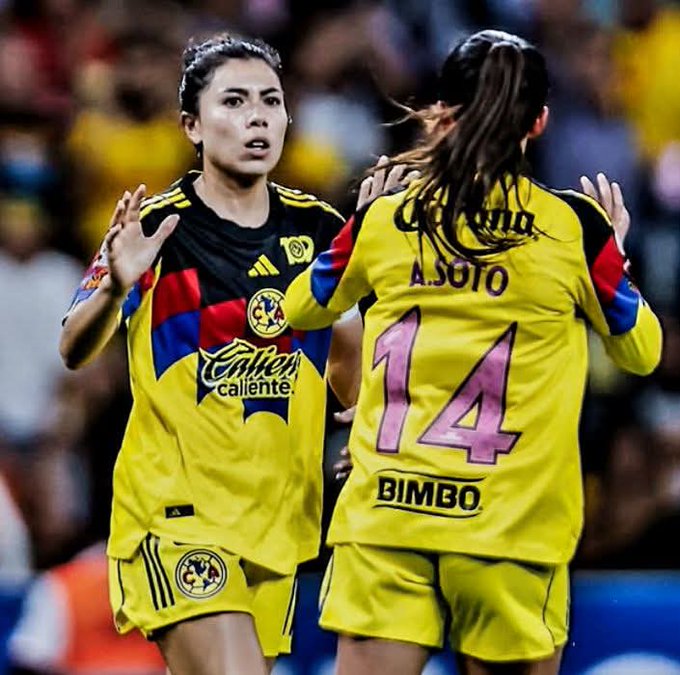 Two female soccer players celebrate a goal on a stadium field, one in a yellow jersey numbered 14 with Asojo on the back and Bimbo sponsor logo, the other in a multicolored jersey with America and Cerveza Caliente sponsor logos, both wearing matching yellow shorts, embracing with arms outstretched amid a blurred crowd in the background.
