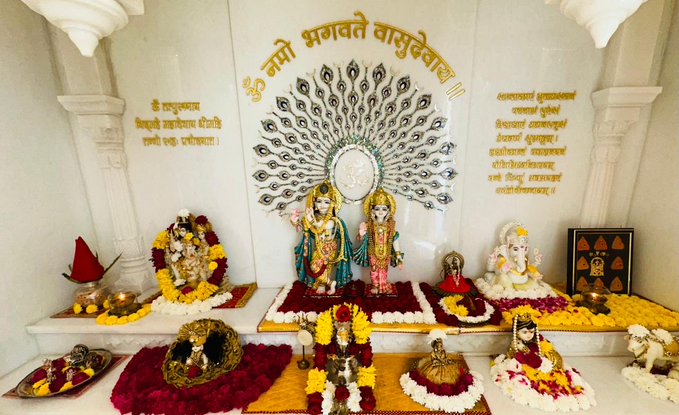 First image shows a statue of young Krishna adorned with gold jewelry, a peacock feather crown, and black curly hair, seated on a golden swing surrounded by red rose petals and yellow marigold flowers on a white marble altar. Second image depicts white marble statues of Radha and Krishna standing side by side on a decorated platform with yellow and red flowers, lamps, and offerings, behind a large peacock motif wall with Devanagari script inscription and an Om symbol.