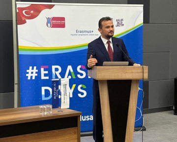 First image shows a man in suit standing at wooden podium gesturing with hand raised speaking into microphone backdrop features blue banner with Erasmus+ logo Turkish flag and text including ErasmusDays event name second image captures large diverse group of people men women in formal casual attire seated standing in spacious lit auditorium with stage Turkish flags tables and chairs third image depicts group of suited men women some in headscarves standing near glass display cases containing medals awards in university corridor with Artuklu University signage fourth image shows group of formally dressed individuals men women around blue draped table with cake holding Erasmus+ plaque banner overhead.