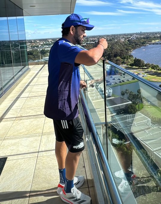 Indian cricketer Rohit Sharma stands on a glass balcony overlooking Perth cityscape with water and buildings under blue sky with clouds. He wears blue cap blue sleeveless shirt black Adidas shorts white Adidas sneakers with blue stripes and wristband while leaning on railing with hand on chin looking outward.