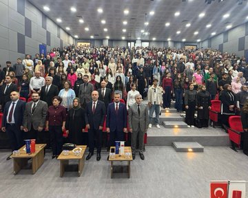 First image shows a man in suit standing at wooden podium gesturing with hand raised speaking into microphone backdrop features blue banner with Erasmus+ logo Turkish flag and text including ErasmusDays event name second image captures large diverse group of people men women in formal casual attire seated standing in spacious lit auditorium with stage Turkish flags tables and chairs third image depicts group of suited men women some in headscarves standing near glass display cases containing medals awards in university corridor with Artuklu University signage fourth image shows group of formally dressed individuals men women around blue draped table with cake holding Erasmus+ plaque banner overhead.