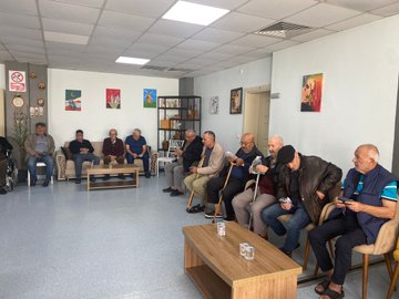 First image shows several men in casual and formal attire seated around wooden tables with glasses of water in a light blue room with shelves, plants, and framed pictures on walls, one man standing and speaking. Second image depicts elderly men some using wheelchairs seated on sofas and chairs in a room with artwork on walls including Turkish flags and abstract paintings, low table with water glasses in center. Third image captures a diverse group of men and women in suits, dresses, and headscarves standing together on a paved area with yellow borders and red carpet leading to an orange building entrance under a portico. Fourth image illustrates women mostly wearing headscarves and long dresses seated around a wooden table with water glasses in a bright room with large windows, plants, and a television on wall.