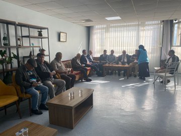 First image shows several men in casual and formal attire seated around wooden tables with glasses of water in a light blue room with shelves, plants, and framed pictures on walls, one man standing and speaking. Second image depicts elderly men some using wheelchairs seated on sofas and chairs in a room with artwork on walls including Turkish flags and abstract paintings, low table with water glasses in center. Third image captures a diverse group of men and women in suits, dresses, and headscarves standing together on a paved area with yellow borders and red carpet leading to an orange building entrance under a portico. Fourth image illustrates women mostly wearing headscarves and long dresses seated around a wooden table with water glasses in a bright room with large windows, plants, and a television on wall.