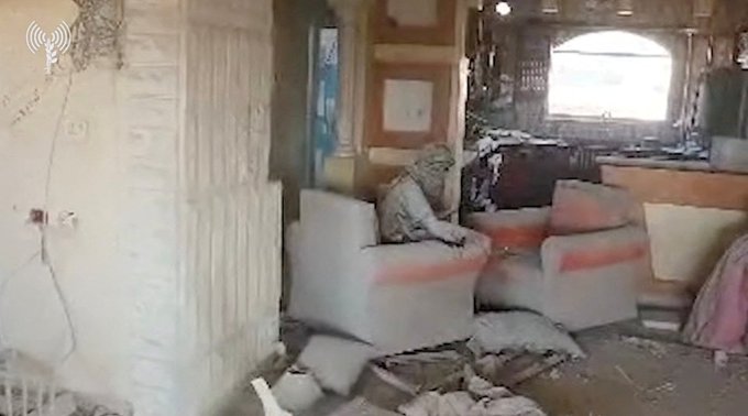 A room with white textured walls partially collapsed, blue door ajar, arched window with bars, wooden shelves, and electrical outlets visible. Two beige armchairs, one with a seated figure in light clothing holding something, the other empty with pink cushion. Brown debris, broken wood, pink fabric, and scattered items on the tiled floor indicate destruction from an explosion.