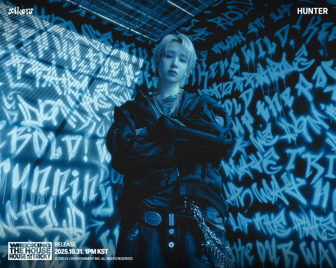 First image shows a young man with blonde hair wearing a black jacket with chains and belts standing with arms crossed in front of a blue graffiti-covered wall featuring text like XIKERS HUNTER and release details for HOUSE OF TRICKY WRECKING THE HOUSE on October 31 2025 at 1PM KST. Second image depicts the same young man in a black puffer jacket with hands in pockets against a similar blue graffiti background with HUNTER text and identical album release information.