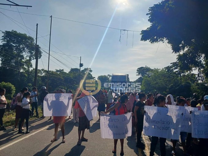 Un grupo de personas de diversas edades, incluyendo mujeres y hombres con ropa casual, sostienen carteles blancos en una carretera pavimentada bajo un cielo soleado con sol brillante. Ãrboles verdes y postes de energÃ­a flanquean la escena, con un bus grande visible al fondo. Algunos carteles muestran texto como Talamanca Escuela, y el logo de Emisoras Unidas aparece en la esquina inferior derecha.