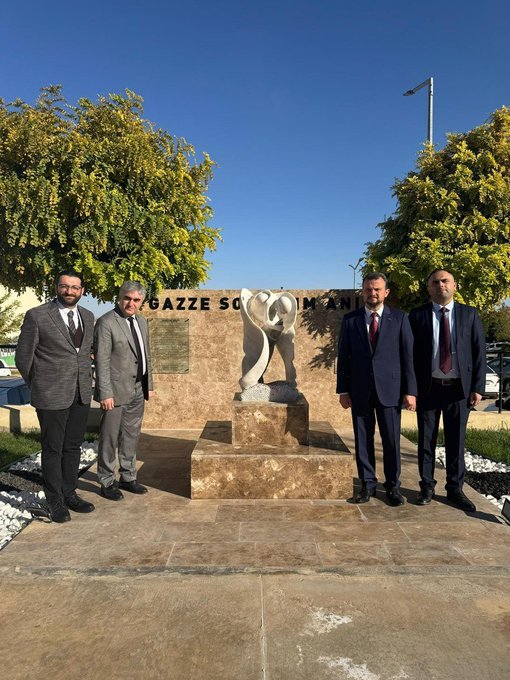 First image shows a woman in white hijab and abaya standing next to a man in dark pinstripe suit, both facing a beige stone wall with black text Gazze Sokağı engraved on a plaque, surrounded by yellow-leaved trees and parked cars on a sunny day. Second image depicts four men in suits, including one in gray and three in dark suits with ties, standing together in front of a beige stone pedestal holding an abstract white sculpted form resembling intertwined shapes or a heart, with Gazze Soykırım Anıtı text on the base, yellow trees and blue sky in background. Third image features four men in suits with ties posing outdoors near a beige building with a large white banner reading Cozum Kudüs Yaşam İttifakı and directional arrow sign for Türkiye Yasin Arat Kardeşliği and Gazze Sokak, under a lamppost and tree.