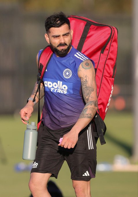 Virat Kohli stands on a green grassy field under a clear sky, wearing a sleeveless purple jersey with white Adidas stripes and Apollo Tyres logo on the chest, black shorts, and white wristband. He has a beard, short dark hair, and visible tattoos on his right arm. He carries a large red duffel bag on his right shoulder, holds a gray reusable water bottle in his right hand, and has a slight frown on his face while looking to the side. In the background, there is a blurred fence and more green field.