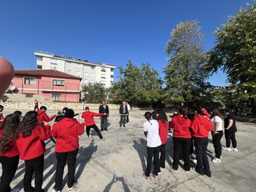 First image shows a large group of young people in red and white sportswear standing together on a paved outdoor area in front of a beige multi-story building with trees and utility poles nearby. Second image depicts the group surrounding two men in suits one extending his arm in a circle formation on the same paved area with the building and trees in background. Third image features a man in a red vest labeled GENC with a microphone standing beside several people in sportswear on the paved area near trees and the building. Fourth image captures young women in red hoodies holding hands in a line performing an activity with jump ropes on the paved courtyard adjacent to the building and basketball hoop.