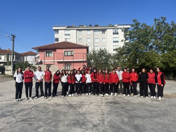 First image shows a large group of young people in red and white sportswear standing together on a paved outdoor area in front of a beige multi-story building with trees and utility poles nearby. Second image depicts the group surrounding two men in suits one extending his arm in a circle formation on the same paved area with the building and trees in background. Third image features a man in a red vest labeled GENC with a microphone standing beside several people in sportswear on the paved area near trees and the building. Fourth image captures young women in red hoodies holding hands in a line performing an activity with jump ropes on the paved courtyard adjacent to the building and basketball hoop.