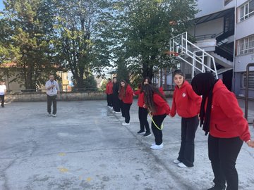 First image shows a large group of young people in red and white sportswear standing together on a paved outdoor area in front of a beige multi-story building with trees and utility poles nearby. Second image depicts the group surrounding two men in suits one extending his arm in a circle formation on the same paved area with the building and trees in background. Third image features a man in a red vest labeled GENC with a microphone standing beside several people in sportswear on the paved area near trees and the building. Fourth image captures young women in red hoodies holding hands in a line performing an activity with jump ropes on the paved courtyard adjacent to the building and basketball hoop.