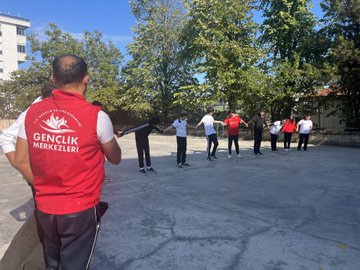 First image shows a large group of young people in red and white sportswear standing together on a paved outdoor area in front of a beige multi-story building with trees and utility poles nearby. Second image depicts the group surrounding two men in suits one extending his arm in a circle formation on the same paved area with the building and trees in background. Third image features a man in a red vest labeled GENC with a microphone standing beside several people in sportswear on the paved area near trees and the building. Fourth image captures young women in red hoodies holding hands in a line performing an activity with jump ropes on the paved courtyard adjacent to the building and basketball hoop.
