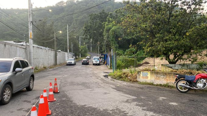 A quiet urban road curves gently with a gray SUV parked on the left side near orange traffic cones arranged in a line. Several other vehicles including white cars and a van are positioned further along the asphalt. A red motorcycle stands on its kickstand beside the cones. A tall white concrete wall with a blue gate encloses the area on one side, backed by dense green trees and rolling hills under a hazy morning sky. Utility poles with wires line the roadside, and a sign is visible on a fence post.